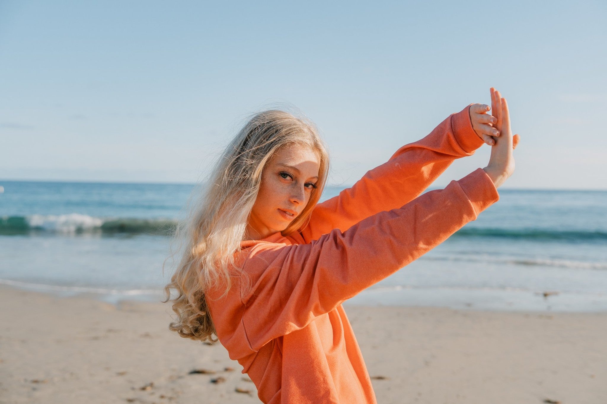 Woman in an orange hoodie taking a posed photo on a beach with ocean in the background