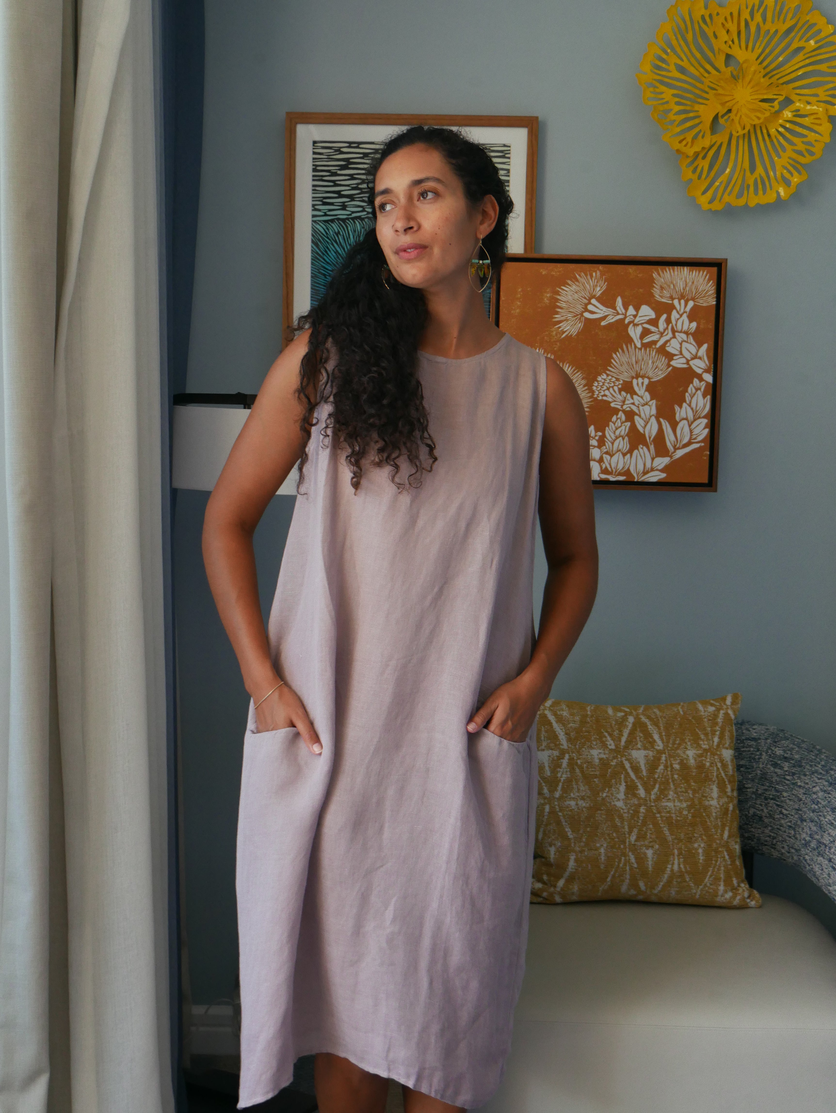 Model in a soft Kahoʻolawe-toned sleeveless linen midi dress with round neckline and pockets, wearing lei, photographed on a Hilo farm.