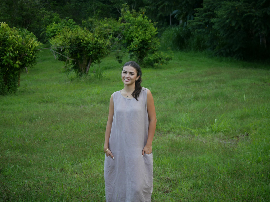 Model in a soft Kahoʻolawe-toned sleeveless linen midi dress with round neckline and pockets, wearing lei, photographed on a Hilo farm.