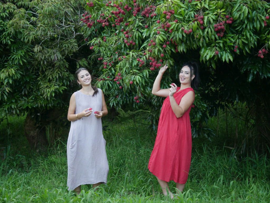 Model in a deep lychee-red cotton A-line dress with V-neckline and pockets, photographed on a Hilo farm.