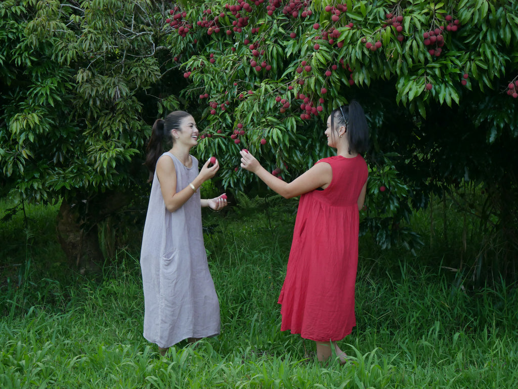 Model in a deep lychee-red cotton A-line dress with V-neckline and pockets, photographed on a Hilo farm.