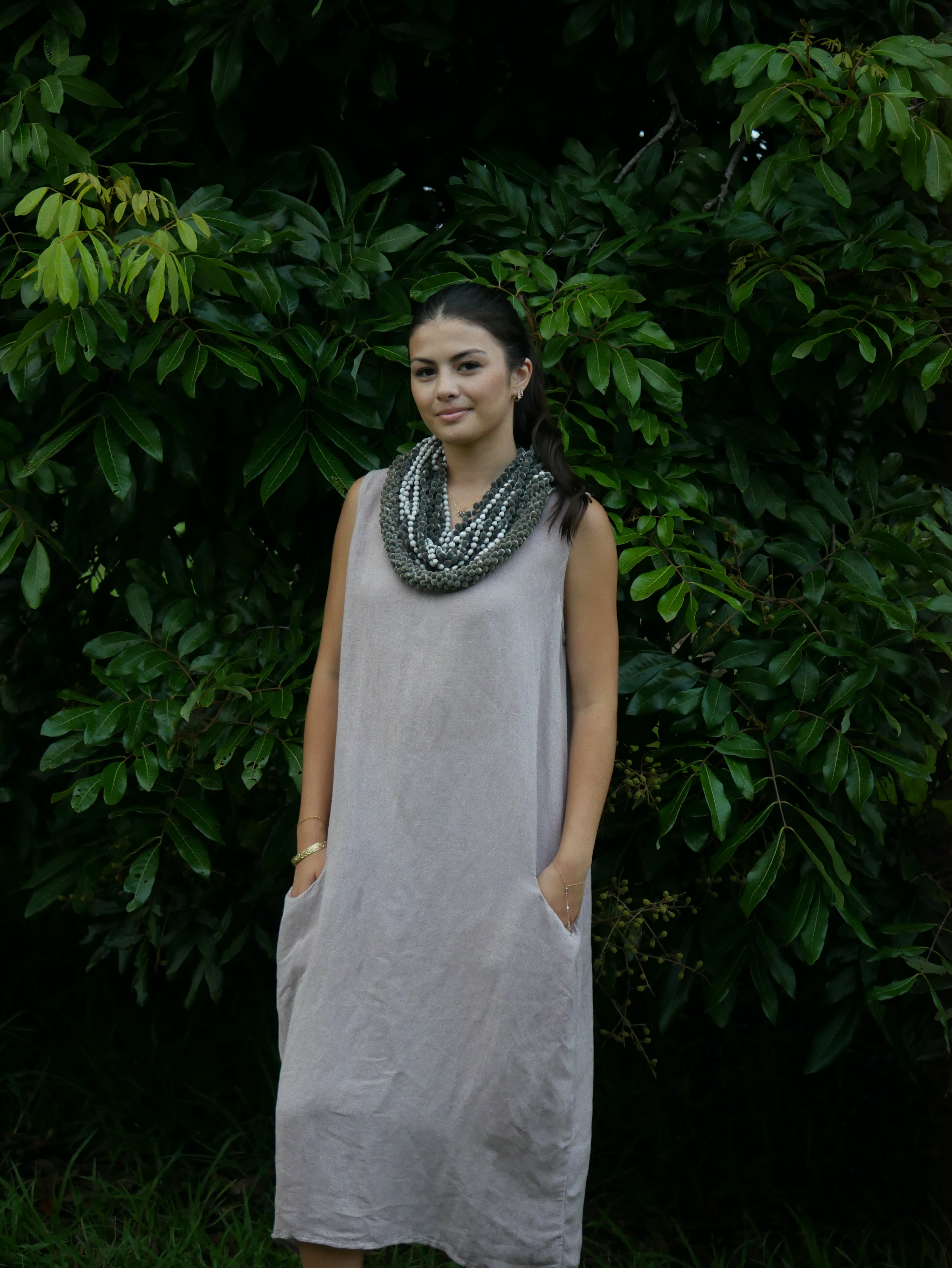 Model in a soft Kahoʻolawe-toned sleeveless linen midi dress with round neckline and pockets, wearing lei, photographed on a Hilo farm.