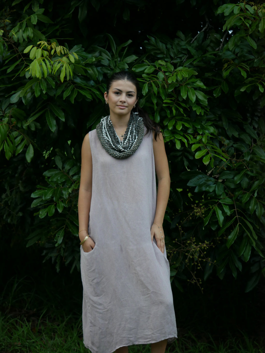 Model in a soft Kahoʻolawe-toned sleeveless linen midi dress with round neckline and pockets, wearing lei, photographed on a Hilo farm.